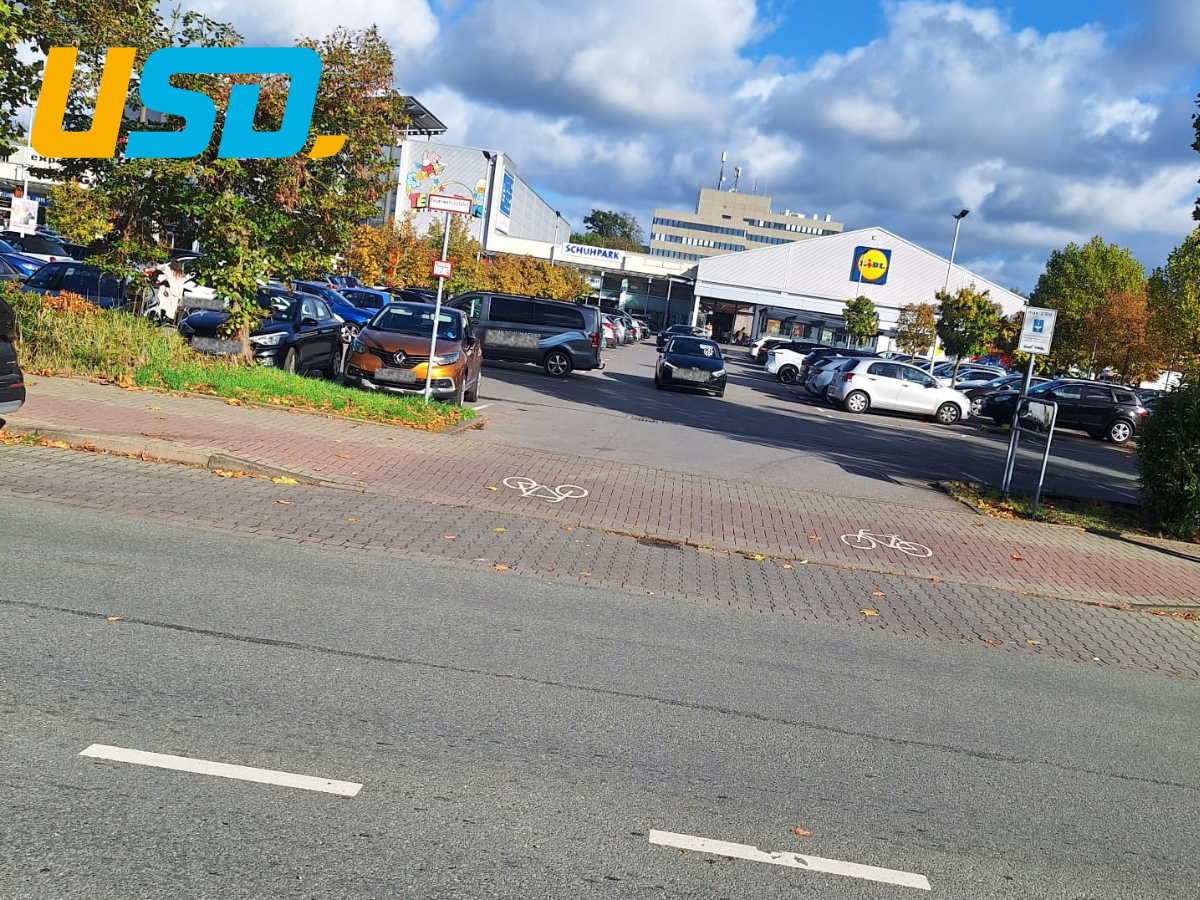 Straßenüberquerung an der Hoffmannstraße mit Radwegmarkierungen und Blick auf einen stark frequentierten Parkplatz vor einem Lidl-Markt.